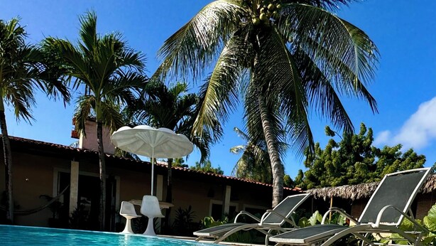 Una piscina cristallina blu con lettini e ombrelloni, circondata da palme e un edificio con tetto di paglia.