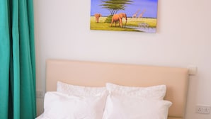Una cama con sábanas blancas, un cabecero color beige y un cuadro que representa una sabana africana sobre ella.