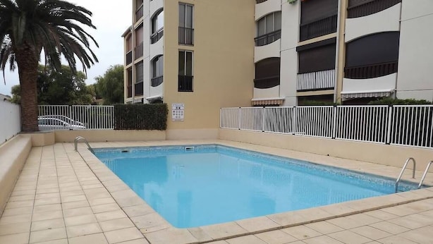 Une piscine dotée d’une terrasse carrelée, entourée d’une clôture blanche et jouxtant un immeuble à plusieurs étages doté de balcons.