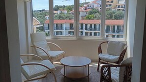 A balcony with wicker chairs and a round table, overlooking a residential area with red-tiled roofs.