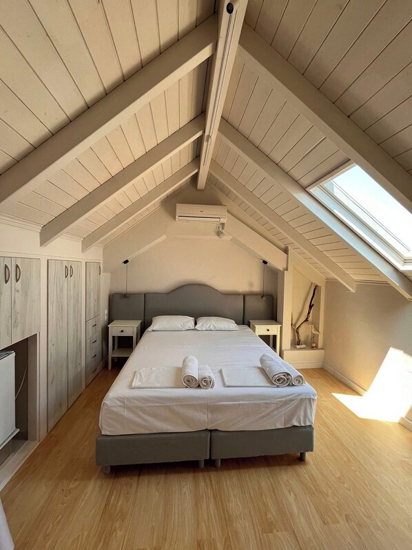 A bedroom with a sloped ceiling, a skylight, a bed with neatly folded towels, a bedside table, and a wardrobe.