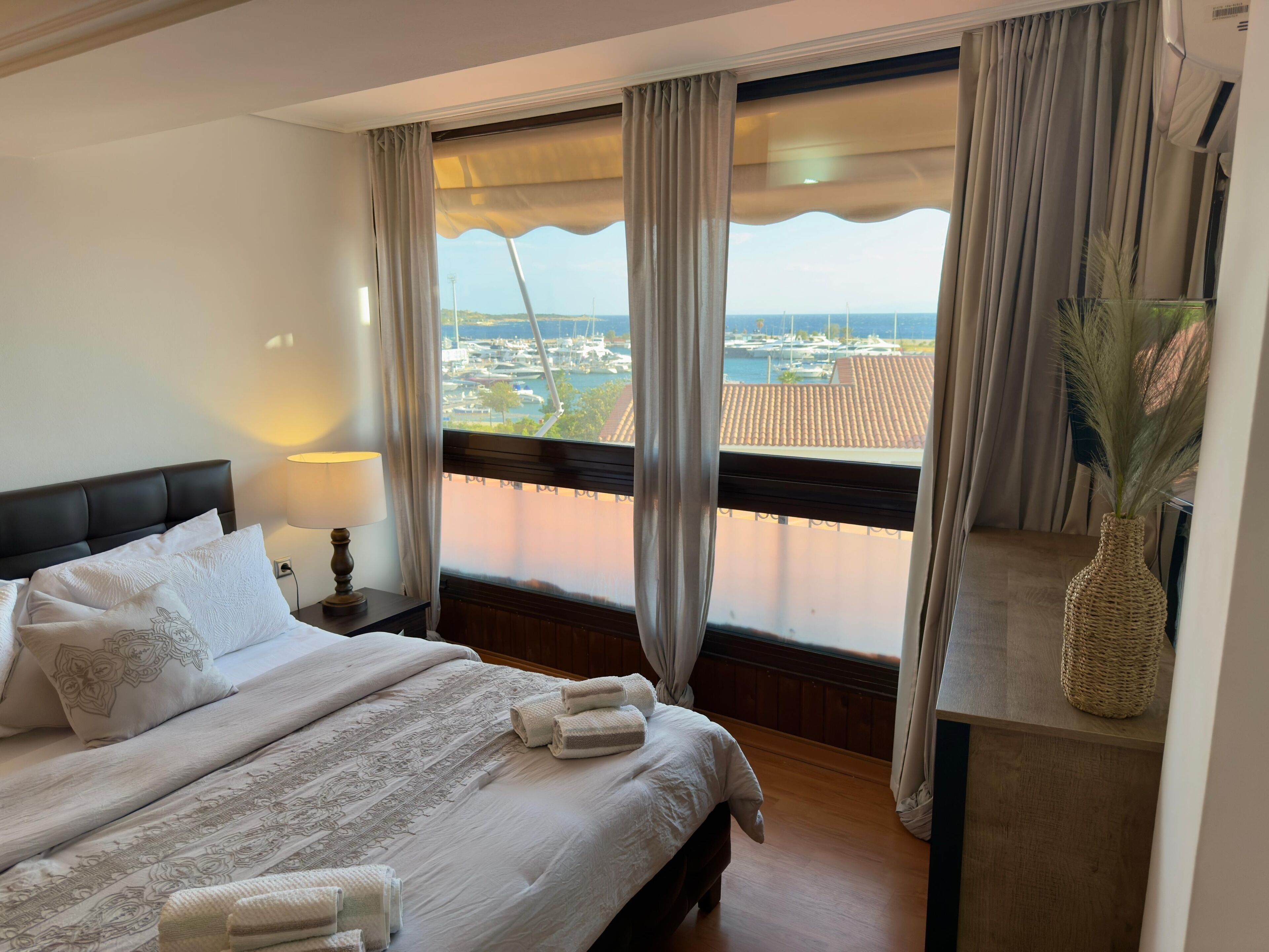A hotel room with a large bed, a nightstand with a lamp, a view of the marina, and a wicker vase on a shelf.