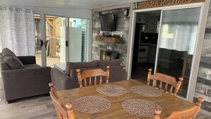 Ein Essbereich mit Holztisch und -stühlen, einem Fernseher über dem Kamin und einer Glastür, die in eine Küche führt.