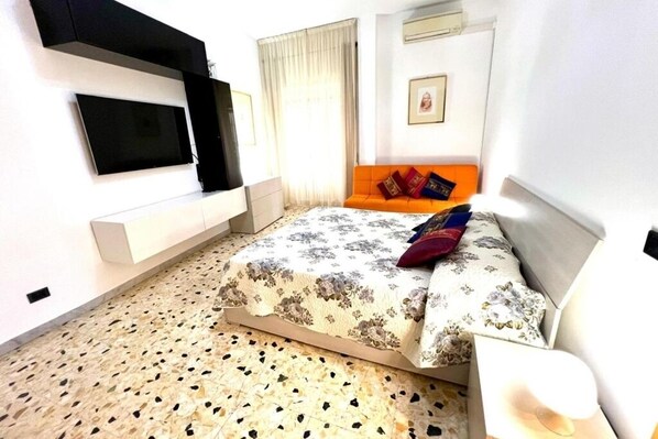 A bedroom with a bed, a TV mounted on the wall, an orange sofa, and a patterned carpet.