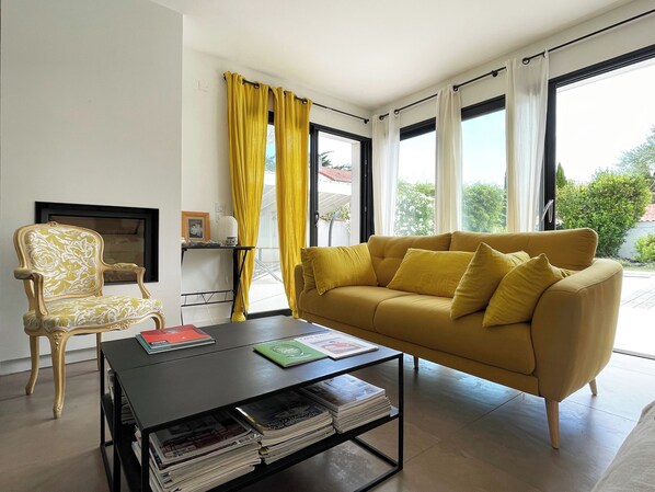 A living room with a yellow sofa, a patterned armchair, a coffee table with magazines, and a view of greenery through large windows.