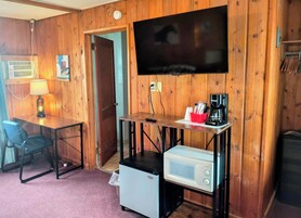 A compact room with wooden walls, a flat-screen TV, a small refrigerator, a coffee maker, and a desk with a chair.