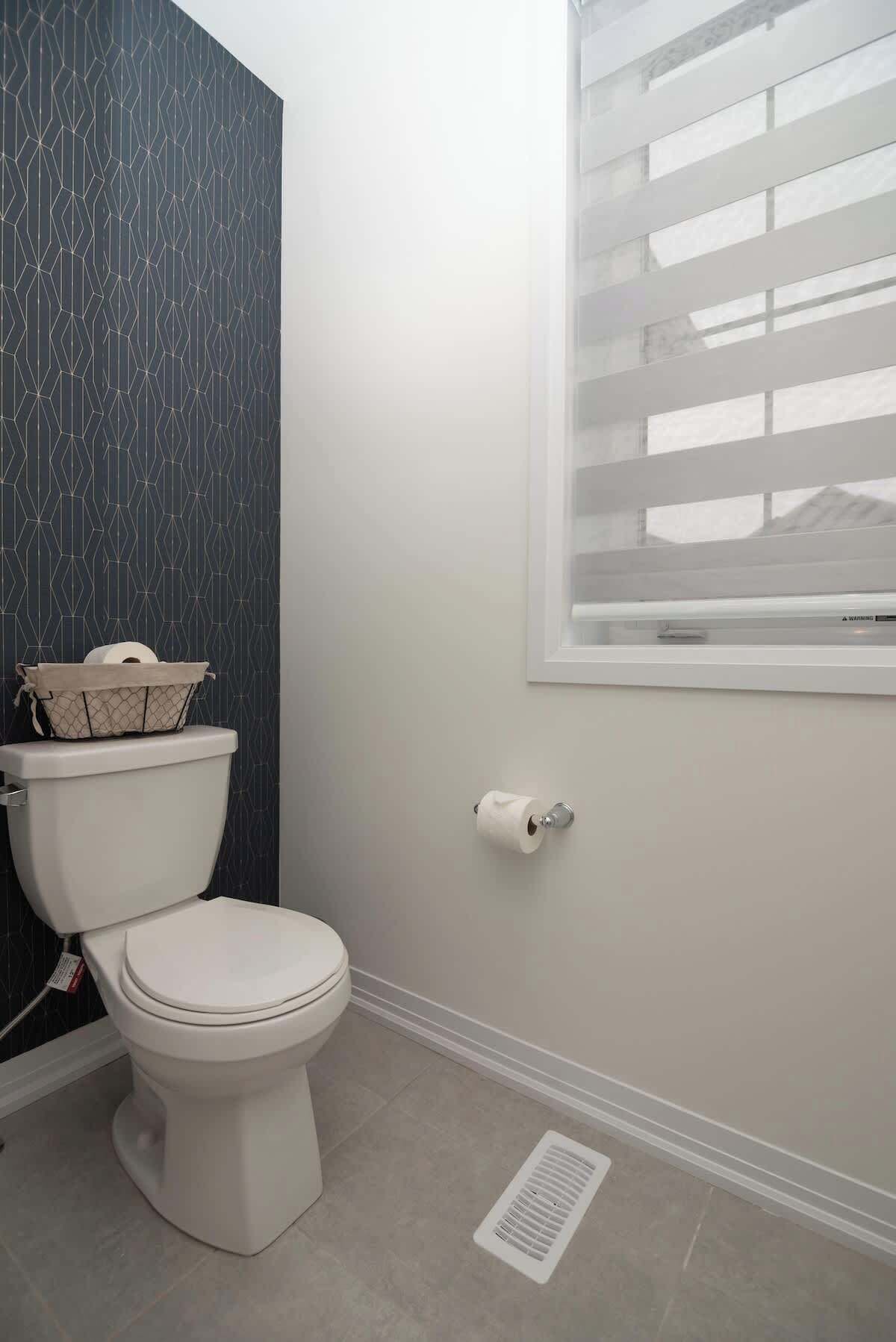A bathroom with a toilet, a window with blinds, a patterned wall, and a floor vent.