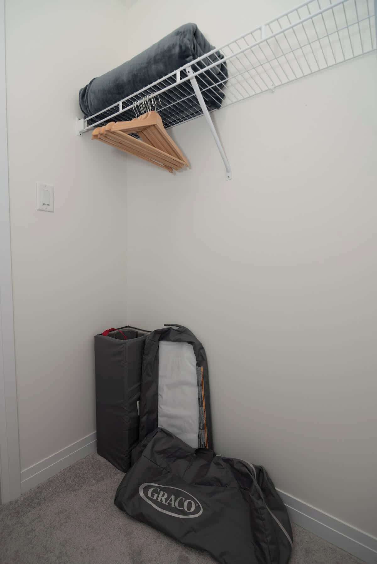 A small room with a white wall, a metal shelf with a folded blanket, a wooden hanger, and a GRACO bag on the floor.