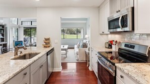 A modern kitchen with white cabinetry, a stainless steel microwave, and a built-in oven. There is a dining area with a table and chairs visible through an open doorway.