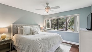 A bedroom with a large bed, a ceiling fan, a window with a view of trees and a car, and a TV on a stand.