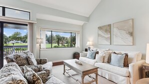 A spacious living room with a large window offering a view of a golf course, a white sofa set, and a patterned armchair.