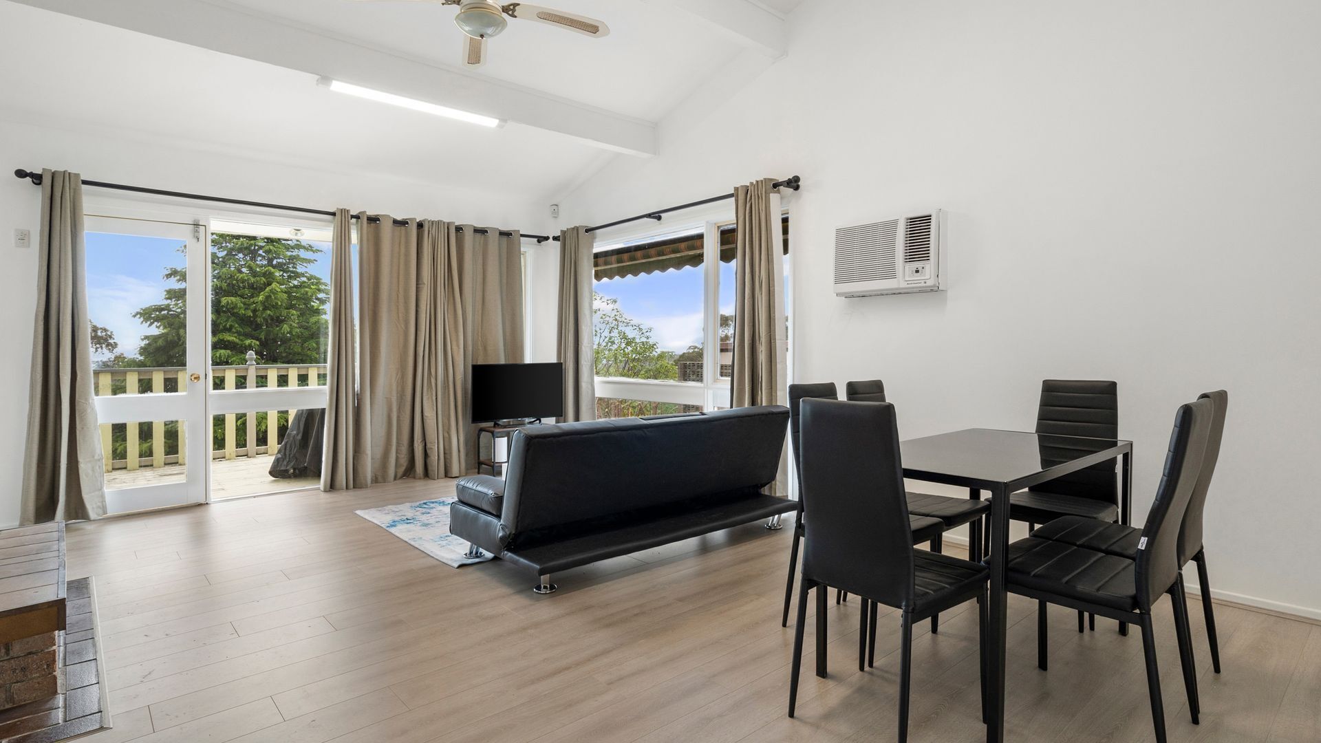 Una sala de estar moderna con sofá, mesa de comedor y sillas. Hay un televisor, un ventilador de techo y una ventana con cortinas.
