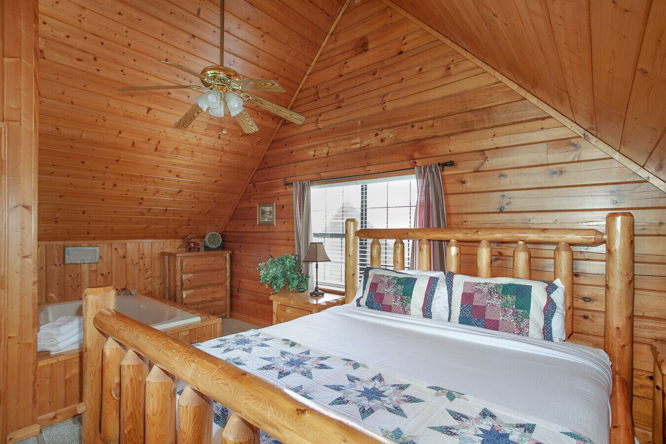 A wooden cabin bedroom with a bed, a desk, a chair, a window, and a ceiling fan.