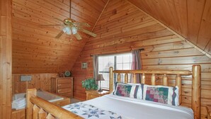 A wooden cabin bedroom with a bed, a desk, a chair, a window, and a ceiling fan.