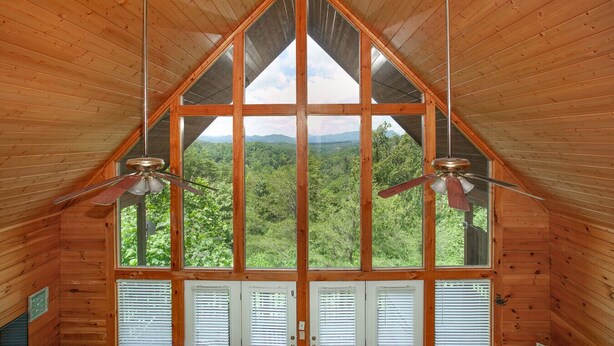 A cabin with a triangular roof, large windows offering a view of a forest, and two ceiling fans.