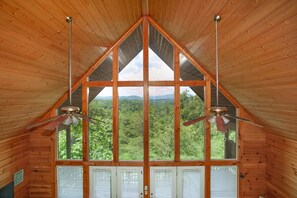 A cabin with a triangular roof, large windows offering a view of a forest, and two ceiling fans.