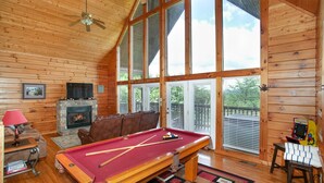 A cozy cabin interior with a pool table, a fireplace, and large windows offering a view of the outdoors.