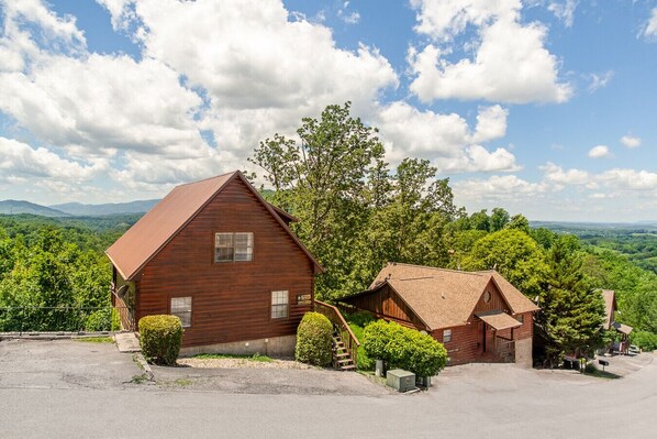 A rustic wooden cabin with a sloped roof, surrounded by lush greenery and a mountainous backdrop.