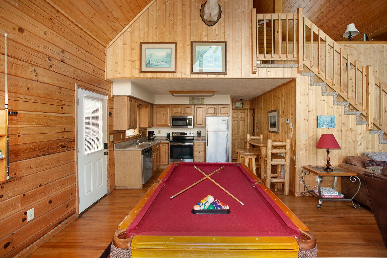 A cozy cabin with a pool table, wooden interior, kitchen, and a staircase leading to a loft.
