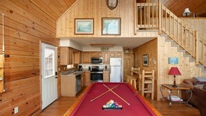 A cozy cabin with a pool table, wooden interior, kitchen, and a staircase leading to a loft.