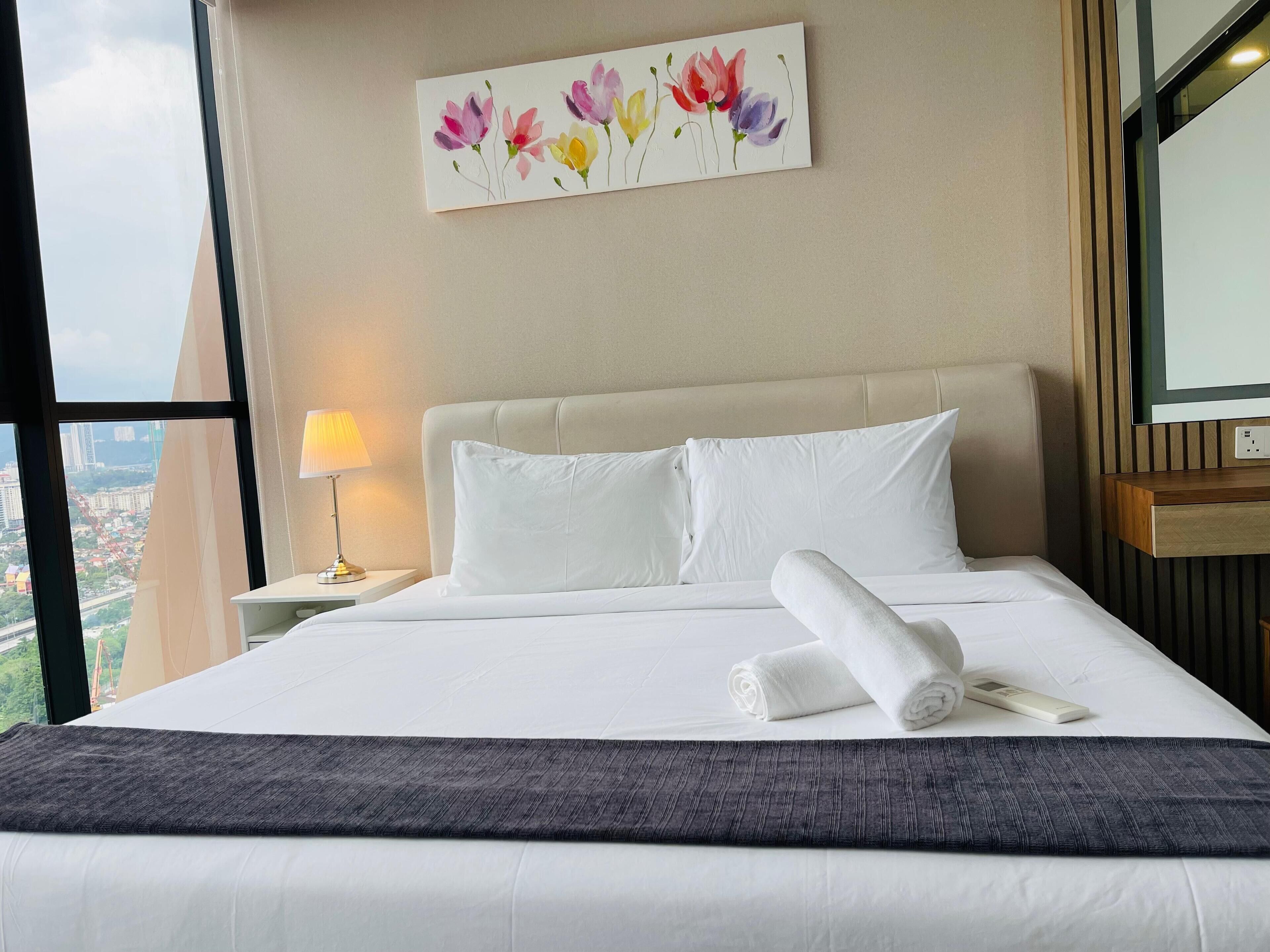 A hotel room with a large bed, white bedding, a view of the city through a window, and a painting of flowers on the wall.