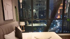 A bedroom with a bed, a nightstand, a lamp, and a view of a cityscape at night. - Scarletz KLCC By Arman (Kuala Lumpur)
