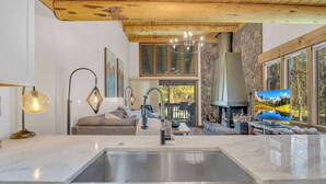 A modern kitchen with a large stainless steel sink, a stone fireplace, and a dining area with a view of the outdoors. - Charming 2-Bedroom Condo with Golf Course Views – Ideal for Families or Couples (Sunriver)