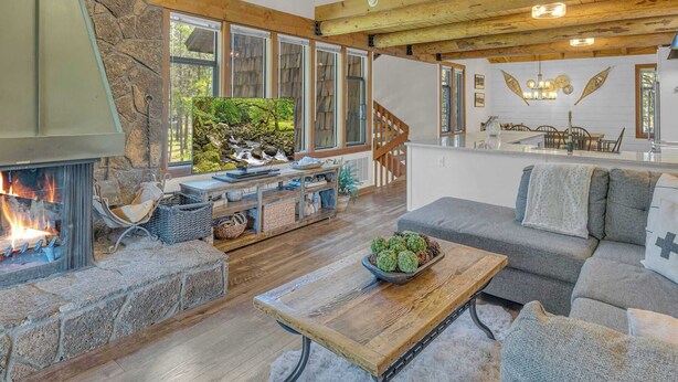 A spacious living room with a stone fireplace, wooden ceiling beams, and a large window overlooking a garden. - Charming 2-Bedroom Condo with Golf Course Views – Ideal for Families or Couples (Sunriver)