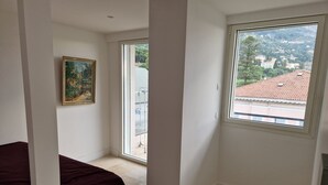 A bedroom with a large window, a painting on the wall, and a bed with a dark cover. - Beautiful sea view, beach 100m away. All comforts. As new (Menton)