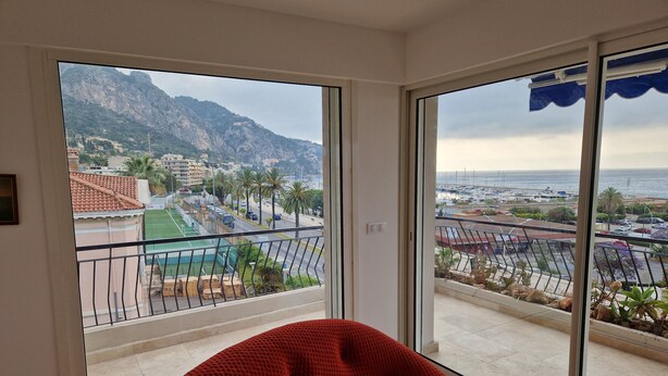 A balcony with a red chair, a view of a mountainous landscape, and a marina with boats. - Beautiful sea view, beach 100m away. All comforts. As new (Menton)