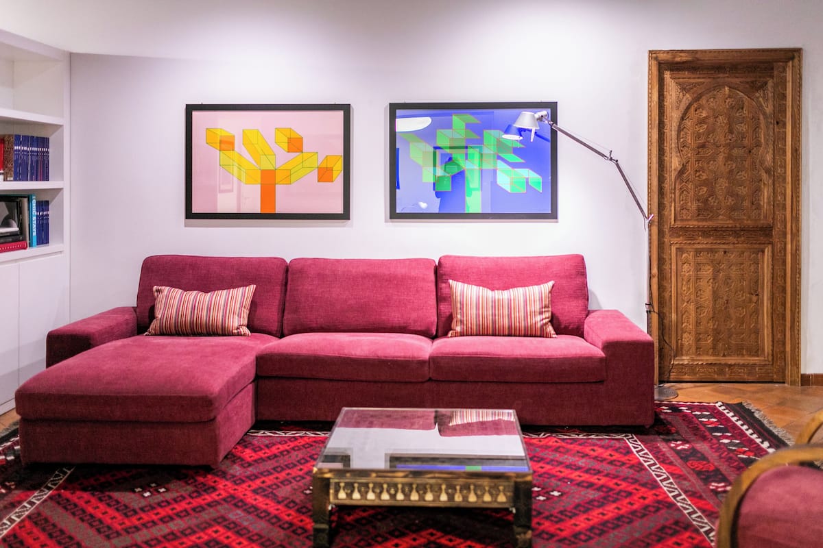 Un salon avec un canapé rouge, deux tableaux abstraits encadrés, une porte en bois et un tapis à motifs.