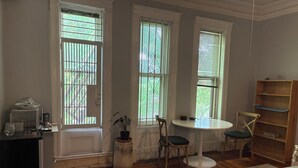 Une pièce comprenant un lit, une petite table ronde avec des chaises, une bibliothèque et trois fenêtres équipées de stores.