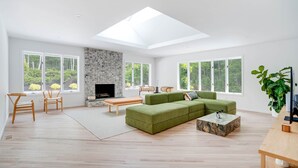 Una sala de estar moderna con un sofá verde, sillas de madera, una chimenea de piedra y ventanas amplias que dan a vistas de vegetación.
