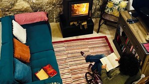En hyggelig stue med en stenvæg, en sort brændeovn, en blå sofa med farverige puder, en gyngestol og en reol.