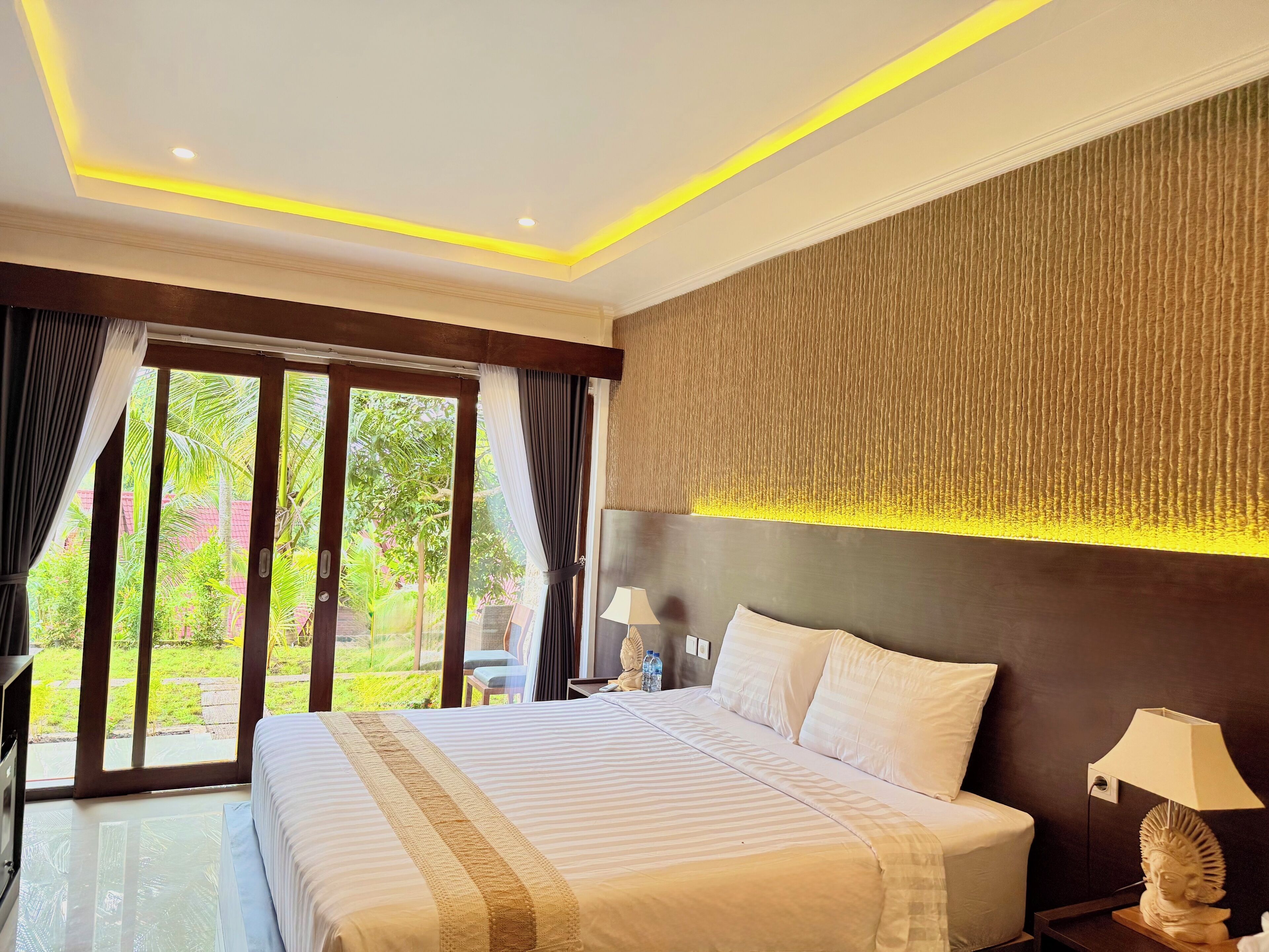 Une chambre d’hôtel moderne, dotée d’un grand lit, d’une table de chevet avec une lampe et offrant une vue sur la verdure à travers les portes vitrées.
