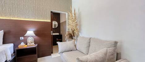 A modern hotel room with a bed, a sofa, a nightstand with a lamp, and a decorative plant.