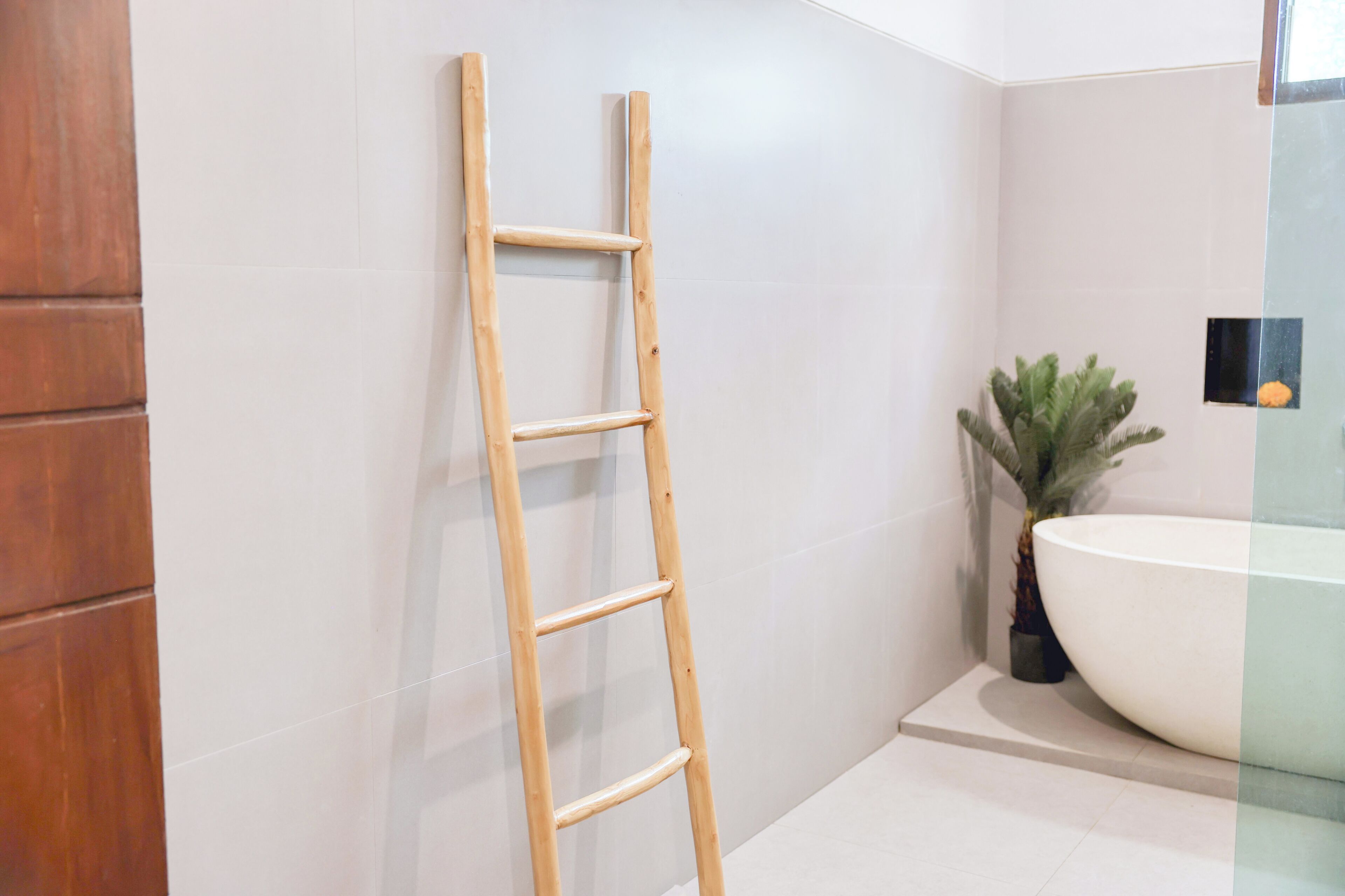 A bathroom with a wooden ladder leaning against a white tiled wall, a large white sink, and a potted plant.