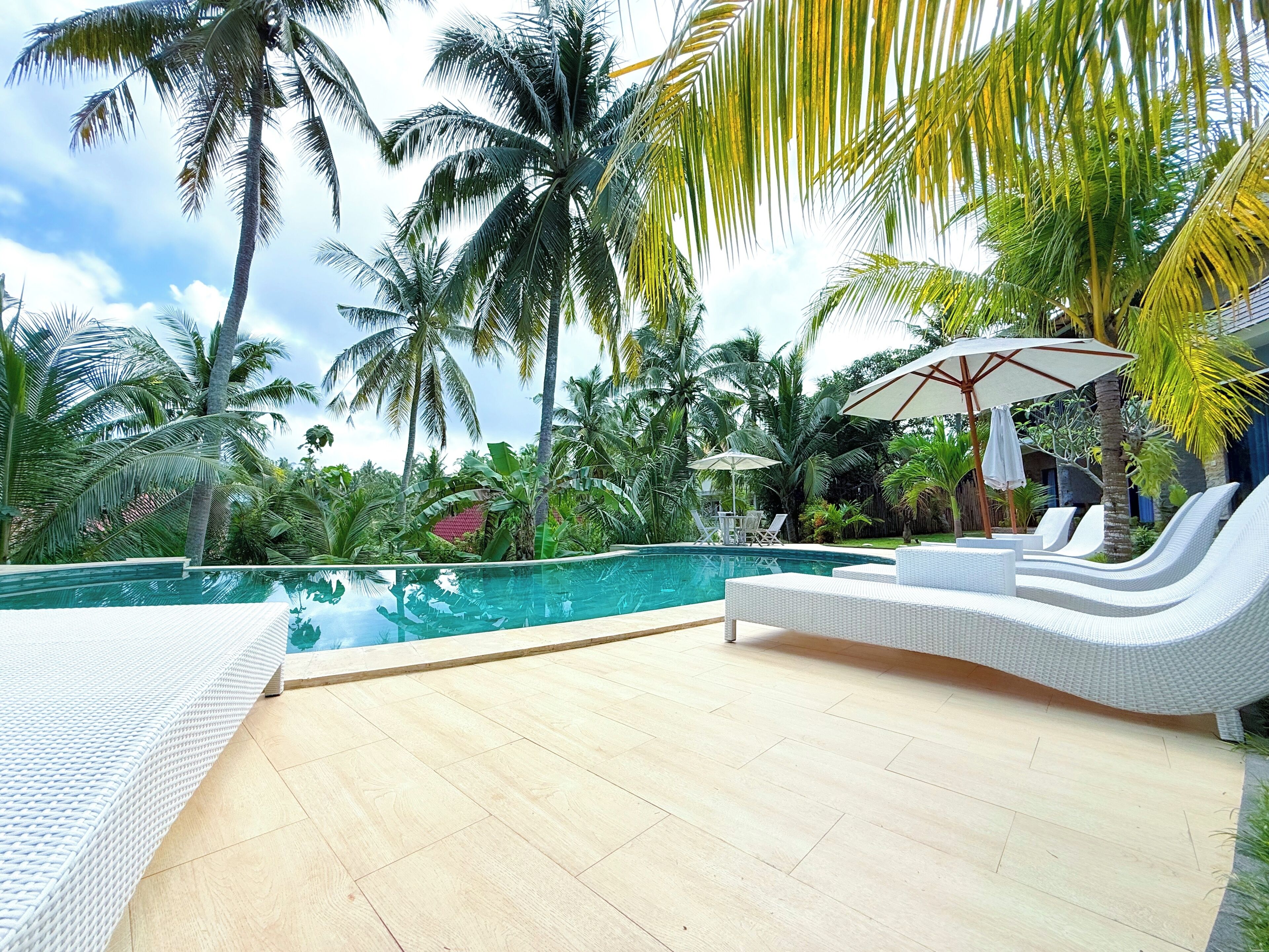 Un espace aménagé au bord de la piscine, avec des fauteuils de relaxation en osier blanc et des parasols, le tout entouré de palmiers et surplombé par un ciel d’un bleu limpide.