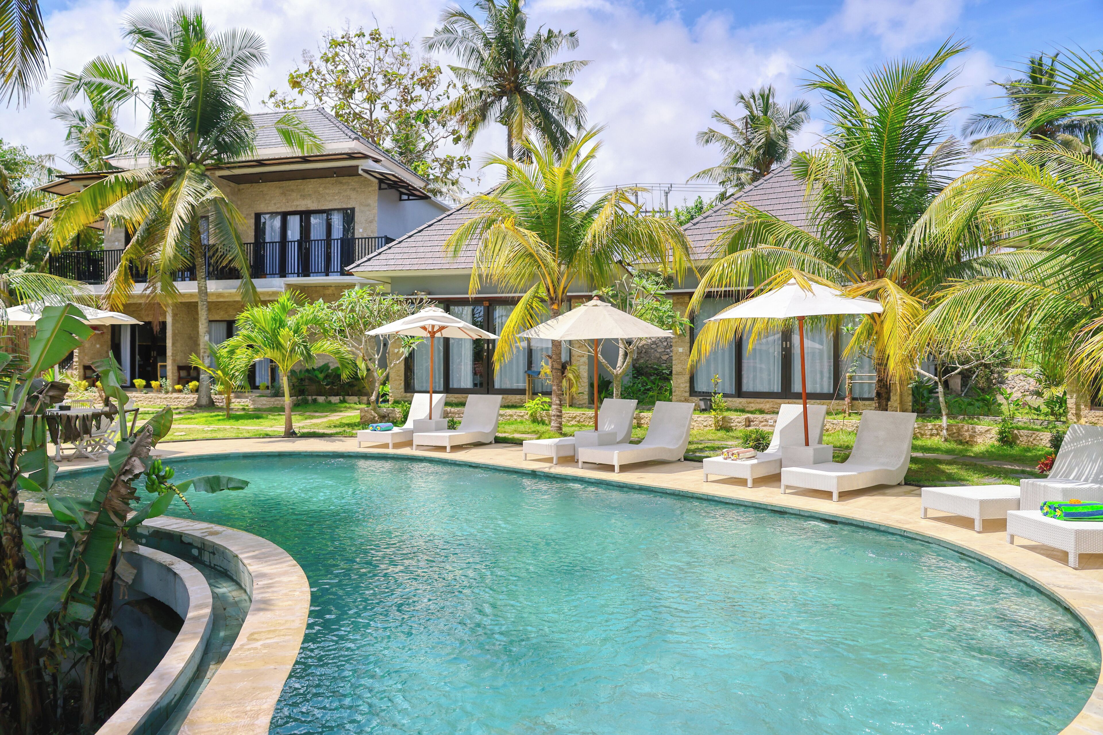 A pool area with lounge chairs and umbrellas, surrounded by palm trees and a building with a sloped roof.