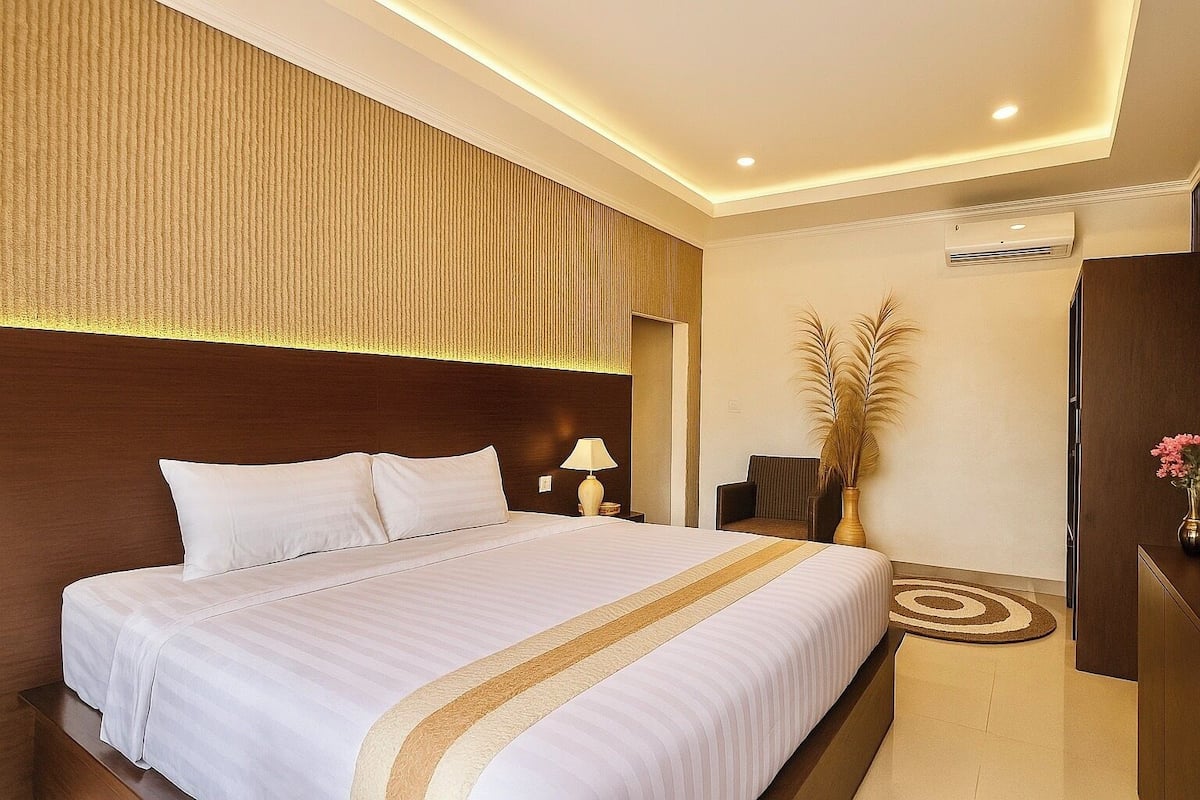 Quarto de hotel com uma cama grande, mesa de cabeceira com abajur, uma cadeira, planta decorativa e guarda-roupa de madeira.