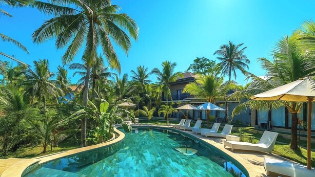 Piscina con sillones de descanso y palmeras, rodeada por un edificio de resort.
