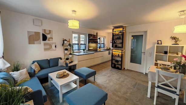 A modern living room with a sofa, ottoman, and coffee table, a kitchen with a central island, and a dining area with a chair and table. - Top modern vacation home family friendly! (Brunnthal)