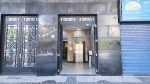 Entrada de un edificio con puertas metálicas ornamentadas, un letrero que dice "Campanella" y una puerta con persianas azules a la derecha.
