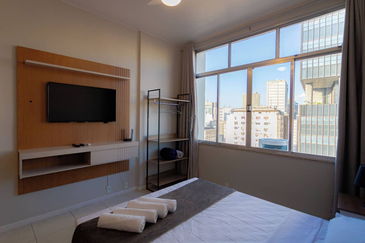 A hotel room with a bed, a TV mounted on the wall, a window with a city view, and a built-in shelf with a hanging rack.