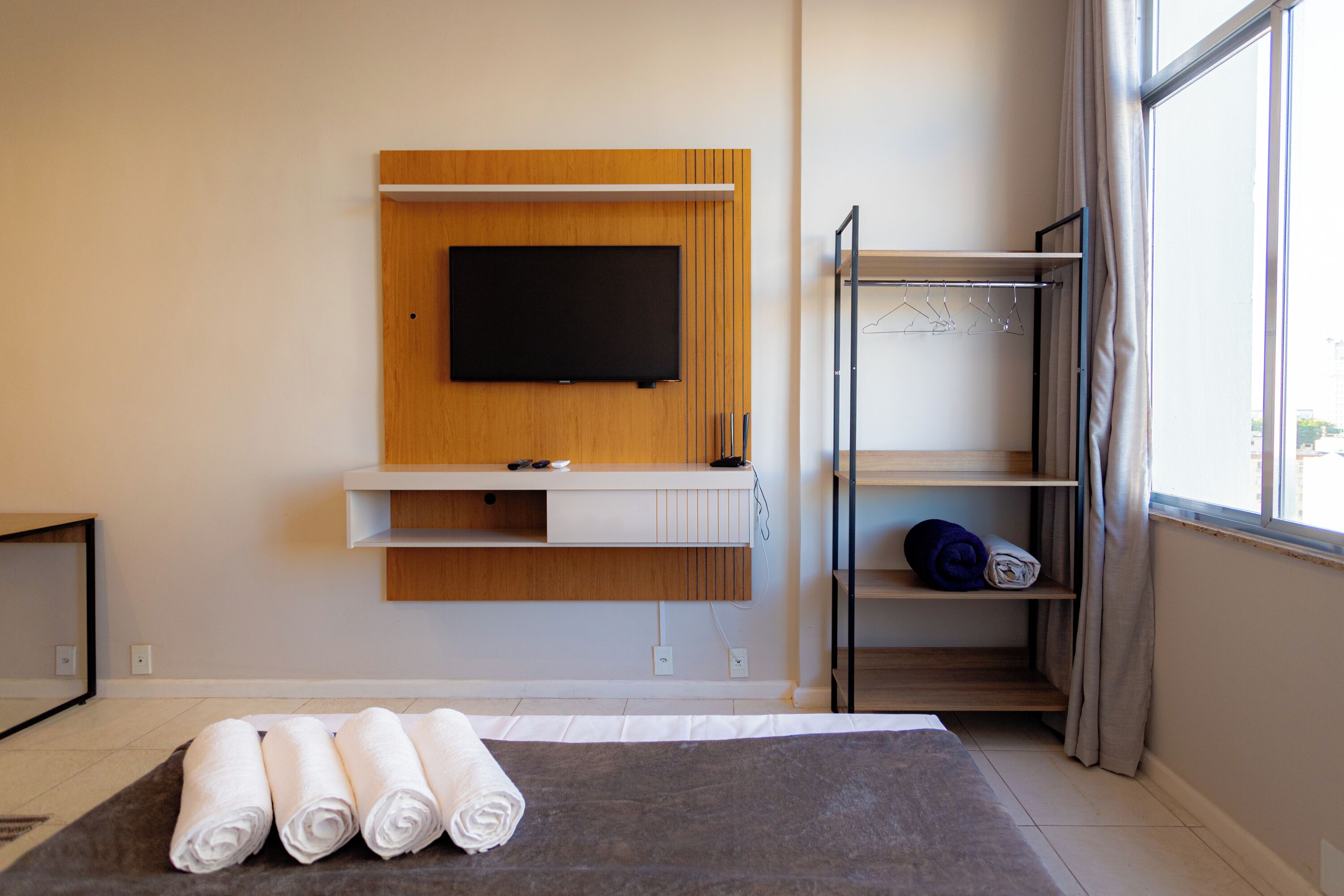 Ein modernes Hotelzimmer mit einem an einer Holzplatte montierten Flachbildfernseher, einer integrierten Ablagefläche mit Handtuch und einem fenster mit Vorhängen.