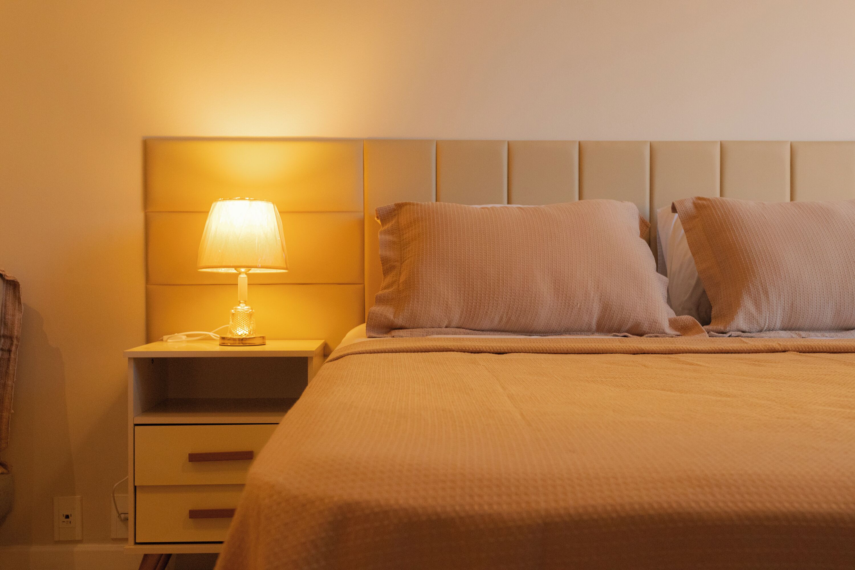 Una cama bien hecha con cabecera, una lámpara de noche y una mesita de noche con cajones.
