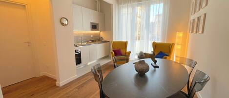 A modern kitchen with white cabinets, a microwave, and a sink. There is a dining area with a round table and chairs, and a window with sheer curtains.