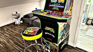 A room with a gaming arcade machine, a stool, a bed, and a window.