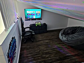 Una habitación con televisor montado en la pared, un sillón de mimbre, un escritorio con computadora y una ventana con persianas verticales.