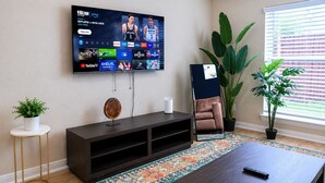 Una sala de estar con televisor de pantalla plana, un mueble de entretenimiento de madera oscura, un sillón de cuero marrón, una pequeña mesa blanca con una planta y una planta grande en maceta.